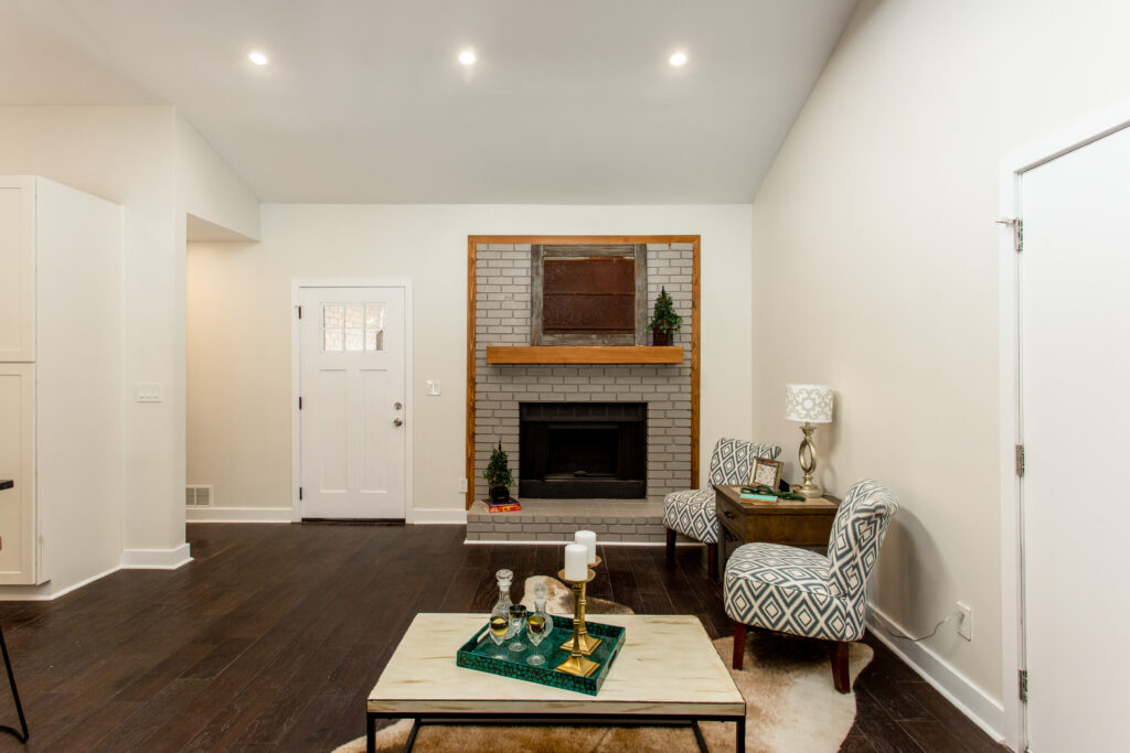 Renovated living room featuring a stone fireplace with wood mantel, vaulted ceiling with exposed beams, white walls, and dark hardwood floors, home renovation by Parsons Mission Construction