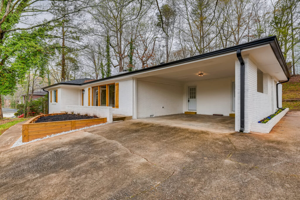 Renovated mid-century modern home featuring white painted brick, wood accent windows, an attached carport, and a raised wooden garden bed with white rock landscaping, exterior renovation by Parsons Mission Construction