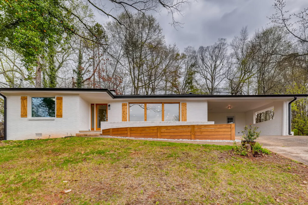 Wide exterior view of a mid-century modern home with white walls, a carport, and surrounding mature trees, exterior renovation by Parsons Mission Construction