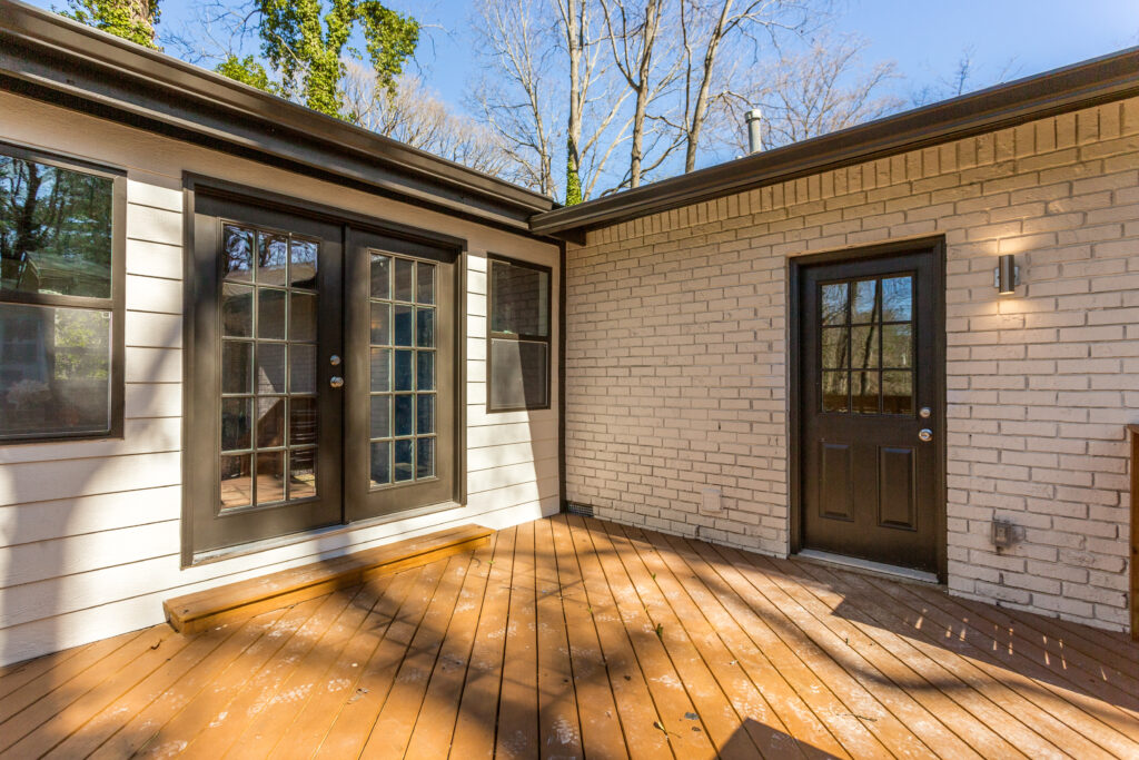 Rear exterior of a home featuring white painted brick, dark French doors, and a wooden deck surrounded by mature trees, exterior renovation by Parsons Mission Construction