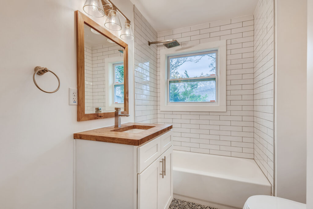Renovated farmhouse bathroom featuring white subway tile walls, a soaking tub, white vanity with wood countertop, wood-framed mirror, and natural light from a window, bathroom remodeling by Parsons Mission Construction