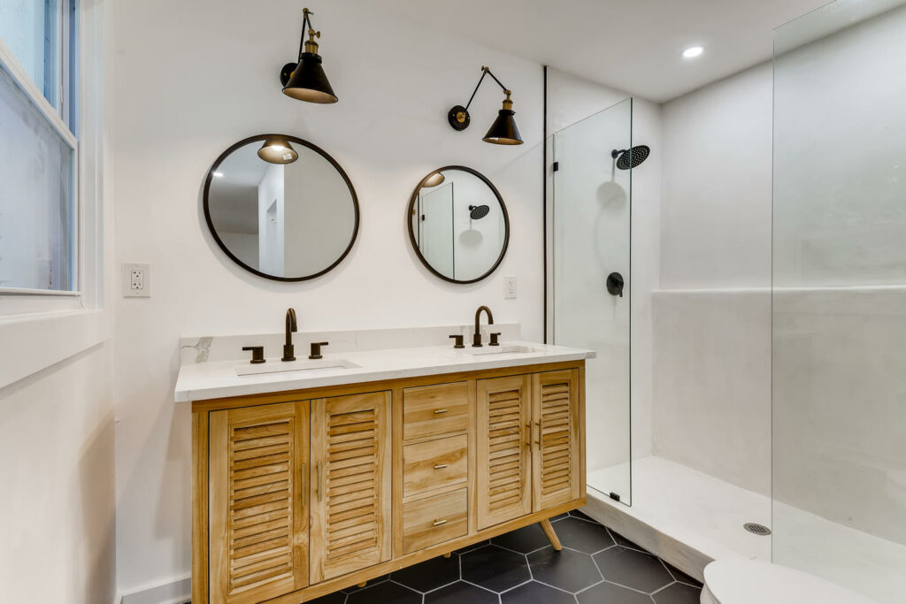 Renovated bathroom featuring a wood vanity with dual sinks, round mirrors, black wall sconces, and a glass enclosed shower, bathroom remodeling by Parsons Mission Construction